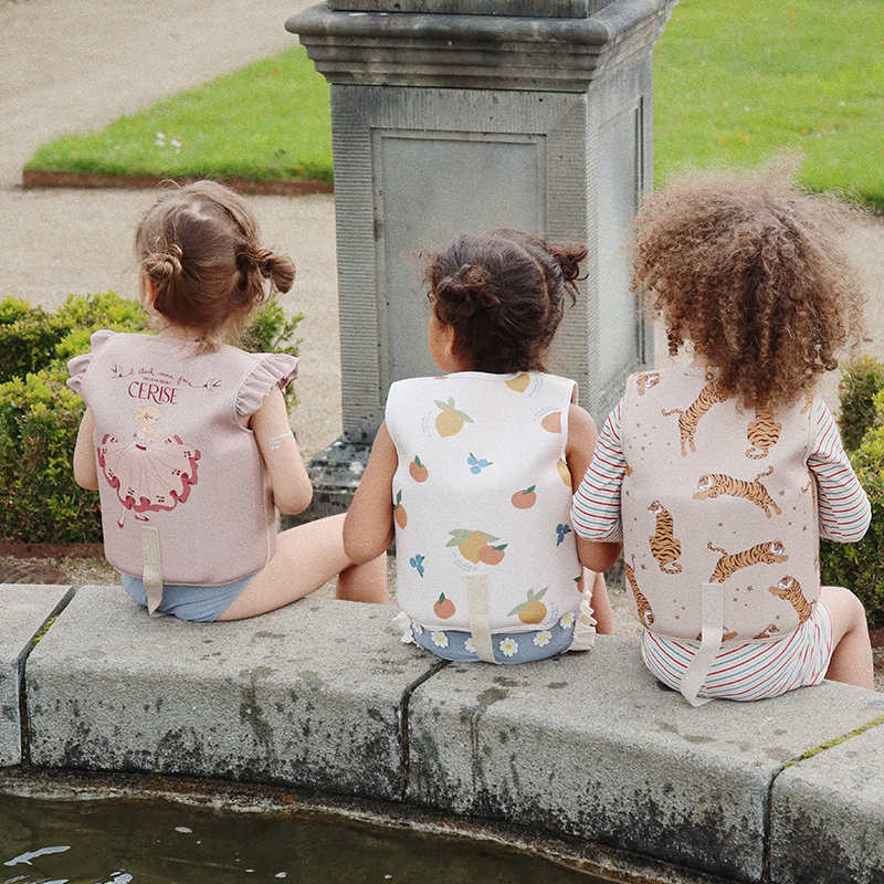 Mädchen sitzen am Brunnen und tragen verschiedene Schwimmwesten mit Prinzessinnenprint, Früchteprint und Tigerprint von Konges Slojd.