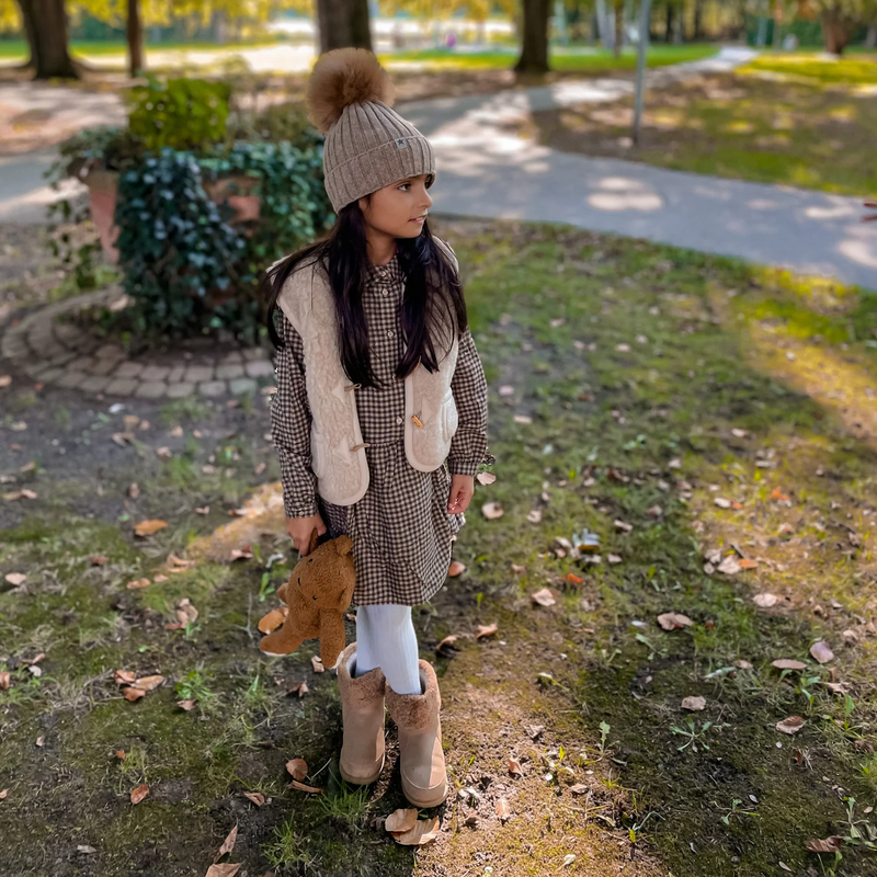 Kind im Park mit Kleid, beige Wollweste der Marke Alwaro, Stiefeln, Mütze und Teddybär in der Hand