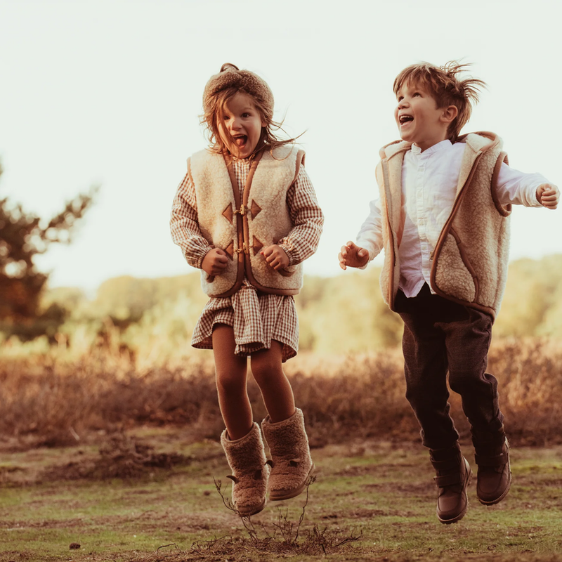 Zwei hüpfende Kinder mit beigen Wollwesten in der Natur