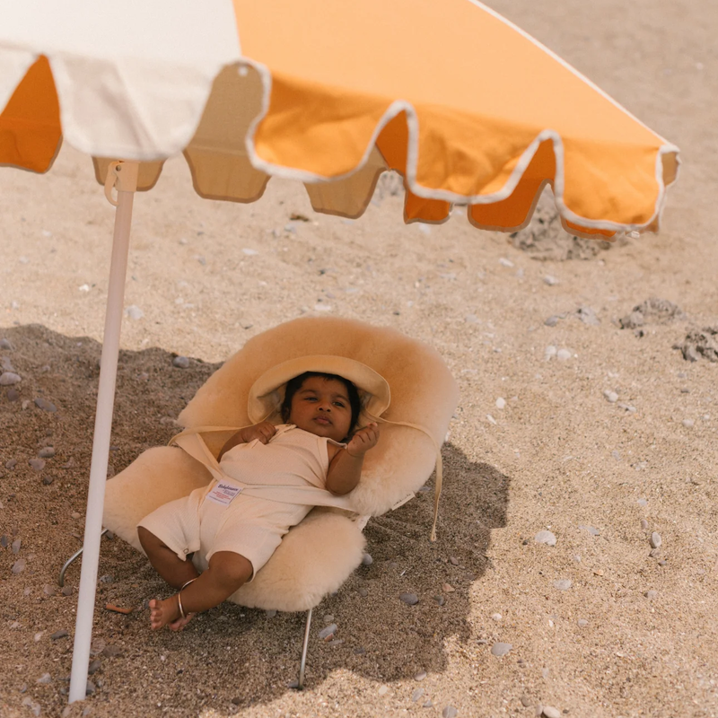 Baby am Strand unter einem Sonnenschirm in Babywippe mit Schaffell-Sitzauflage in Beige