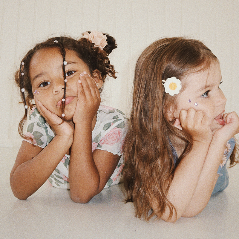 2 Mädchen mit niedlichen Haarfrisuren posieren für ein Foto miteinander. Das Mädchen auf der rechten Seite trägt die Haarclips mit Gänseblümchen von Konges Slojd im Haar.