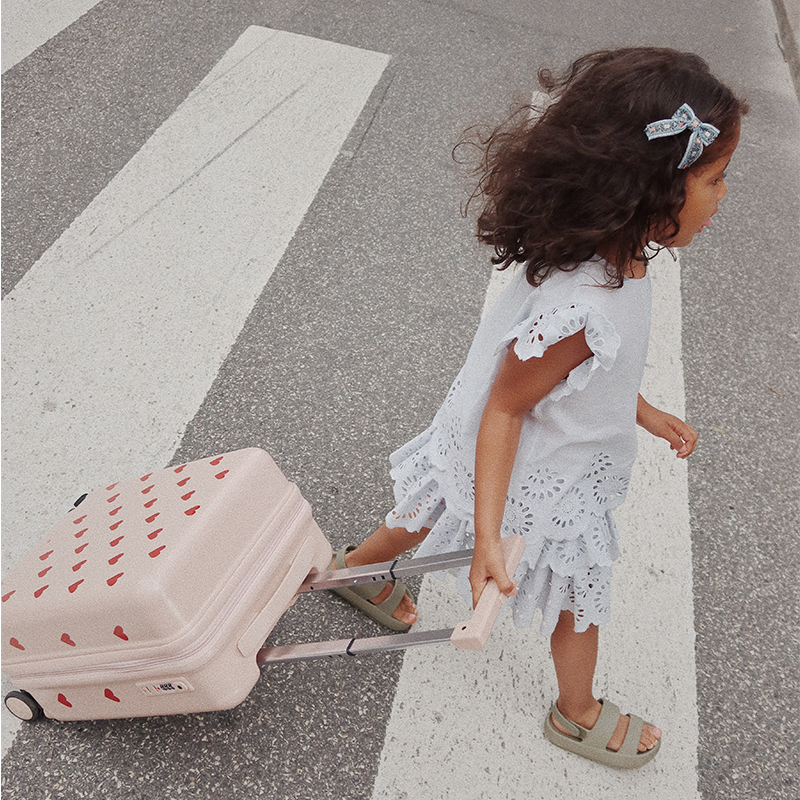 Mädchen zieht einen Reisekoffer in Rosa für Kinder mit Herzchenmuster von Konges Slojd über die Strasse.