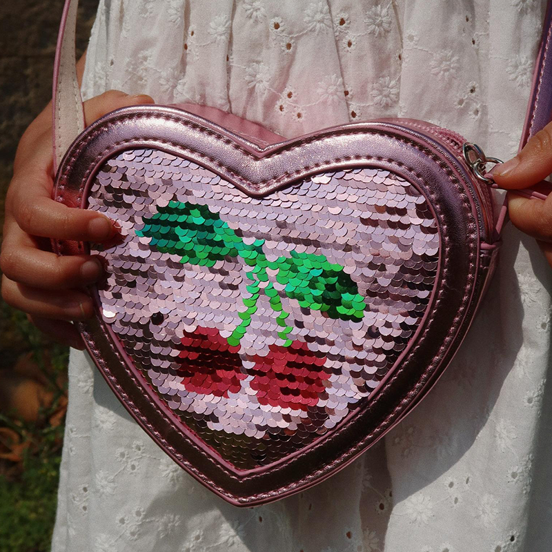 Nahaufnahme von der Umhängetasche in Herzform mit Pailletten in Rosa von Konges Slojd.  Kind hält die Tasche vor dem Bauch.