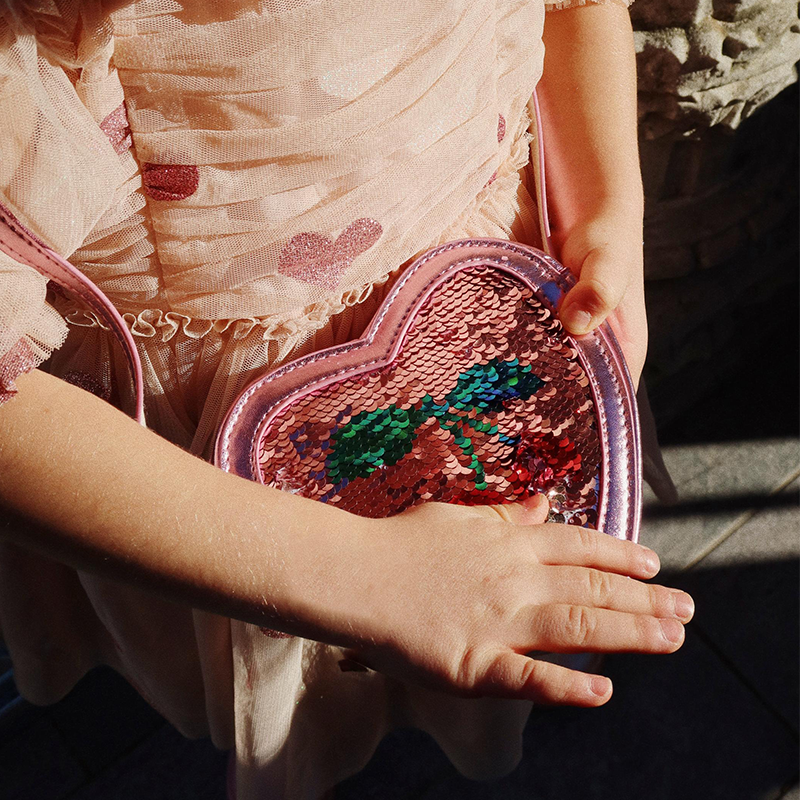 Kinderhände spielen in der Sonne mit der Umhängetasche in Herzform mit Pailletten in Rosa von Konges Slojd. 