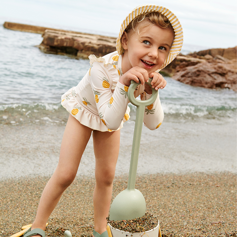 Mädchen am Strand spielt mit einen Eimer und einer Schaufel und trägt den Liewood Badeanzug mit Ananasmuster mit Volants an den Hüften und den Schultern. 