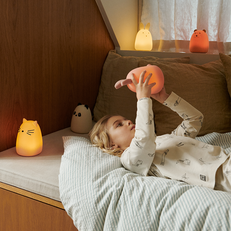 Mädchen liegt im Bett, um sie herum sind verschiedene Nachtlichter. Das Mädchen hält das Nachtlicht in Hasenform in Rosa von Liewood in der Hand.