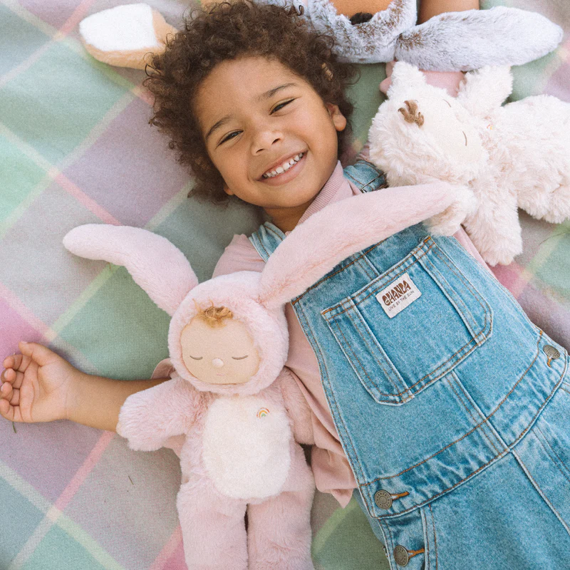 Kind in Jeanslatzhosen liegt mit 3 Dinkum Puppen im flauschigen Hasenkostüm auf einer Decke.