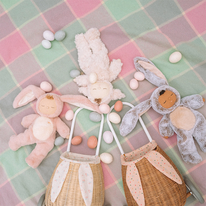 Kuschelige Dinkum Puppen im Hasen- und Lammköstum auf einer pastellfarbenen Decke mit bunten Eiern und Rattankörben um sie herum.