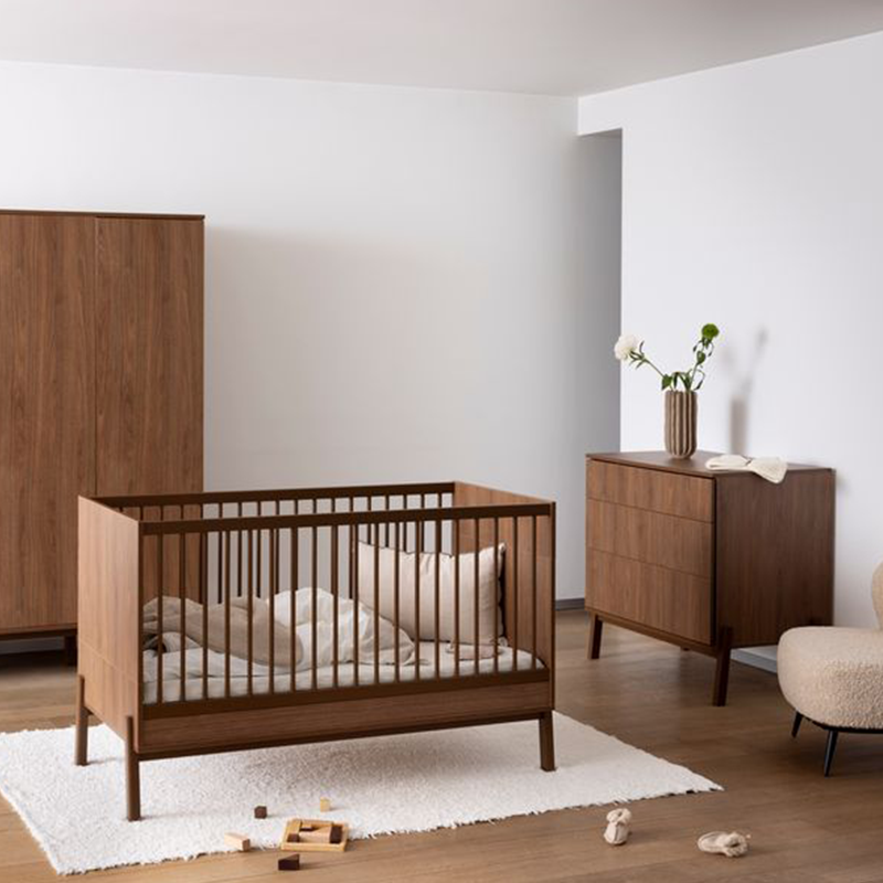 Babybett in Kastanienbraun ohne Matratze von Quax steht in einem Kinderzimmer auf einem weissen Teppich. Im Hintergrund steht ein Schrank und eine Kommode.
