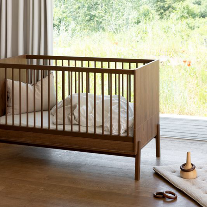 Babybett in Kastanienbraun mit Matratze, Kissen und Decke in Weiss von Quax steht vor einem Fenster.