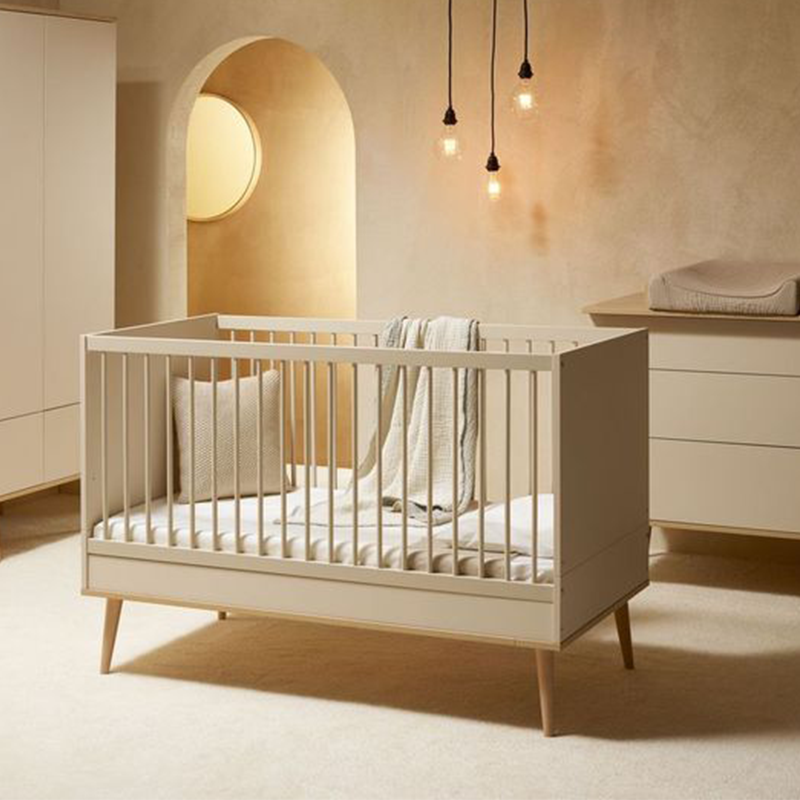 Babybett in Clay mit Beinen aus massiver Buche von Quax in einem Kinderzimmer mit Wickelkommode und Schrank im Hintergrund.