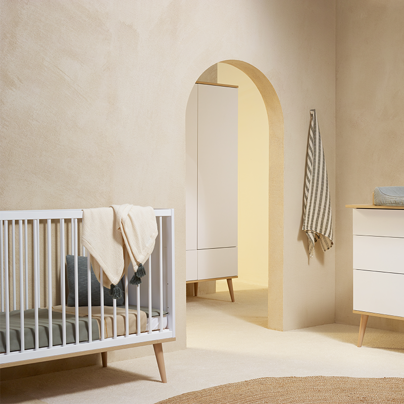 Babybett in weiss mit Beinen aus Eiche von Quax steht in einem Kinderzimmer. Gegenüber vom Bett steht ein passender Wickeltisch.