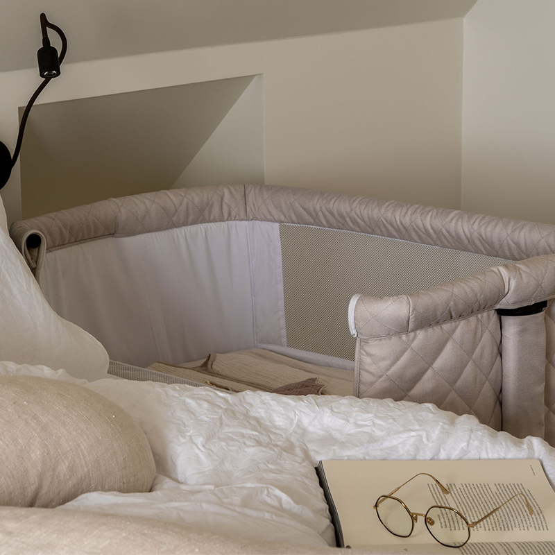 Beistellbett für Babys  in Beige mit schwarzem Gestell steh neben dem Elternbett.