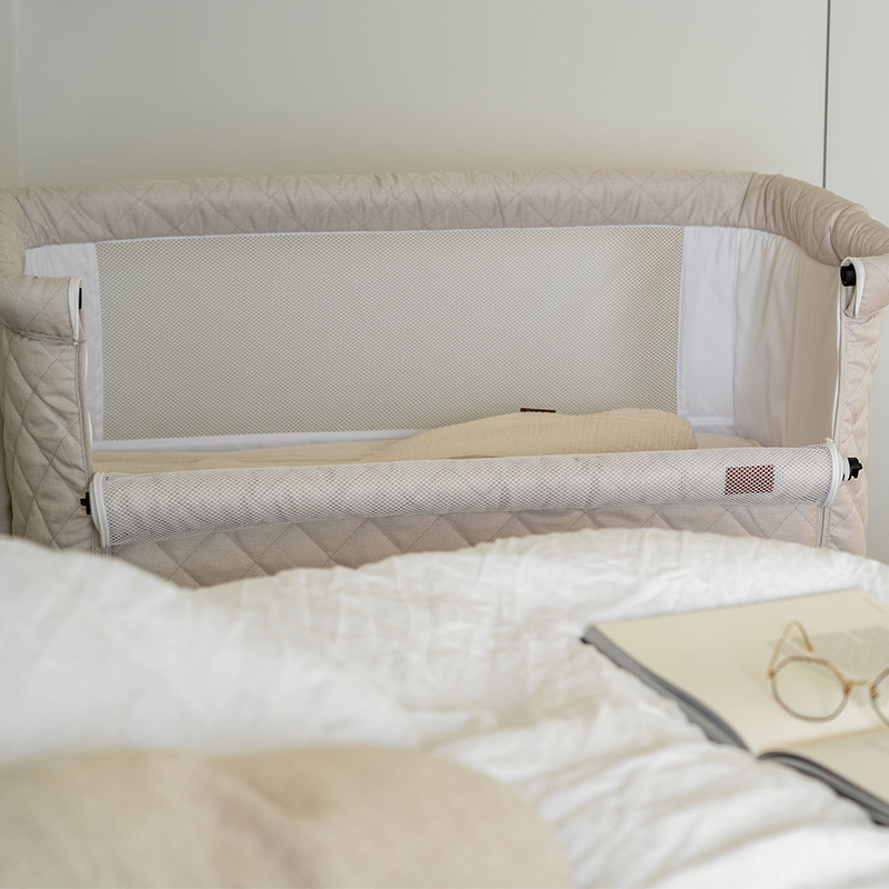 Beistellbett für Babys  in Beige mit schwarzem Gestell von Quax mit offener Seite steht neben Elternbett.