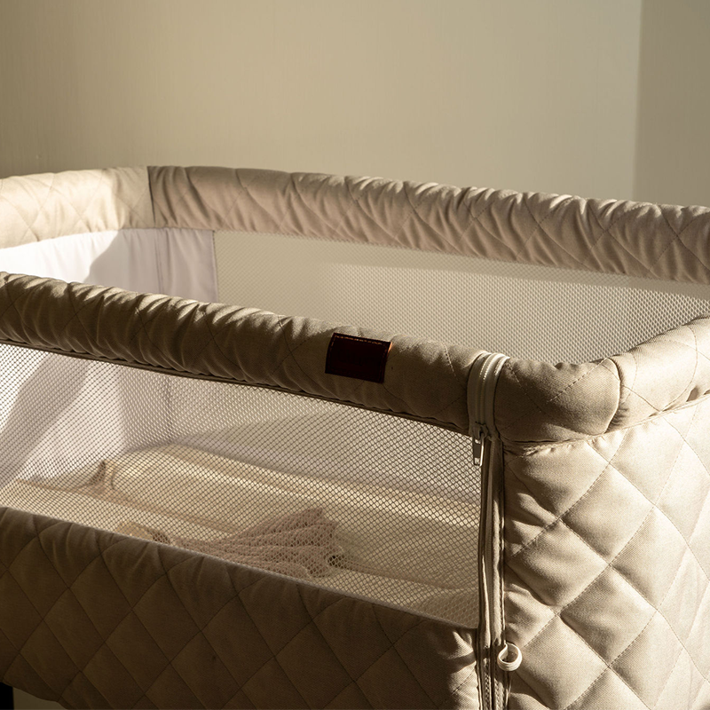 Beistellbett für Babys  in Beige mit schwarzem Gestell von Quax steht in der Ecke eines Zimmers. 
