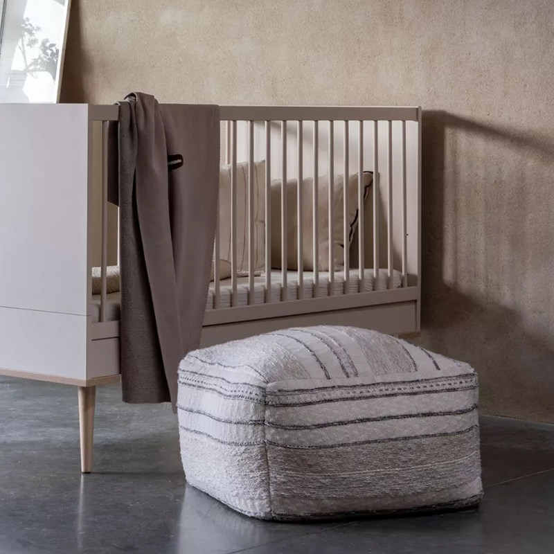 Pouf in Beige mit feinem schwarzen Strickmuster von Quax neben einem Babybett.