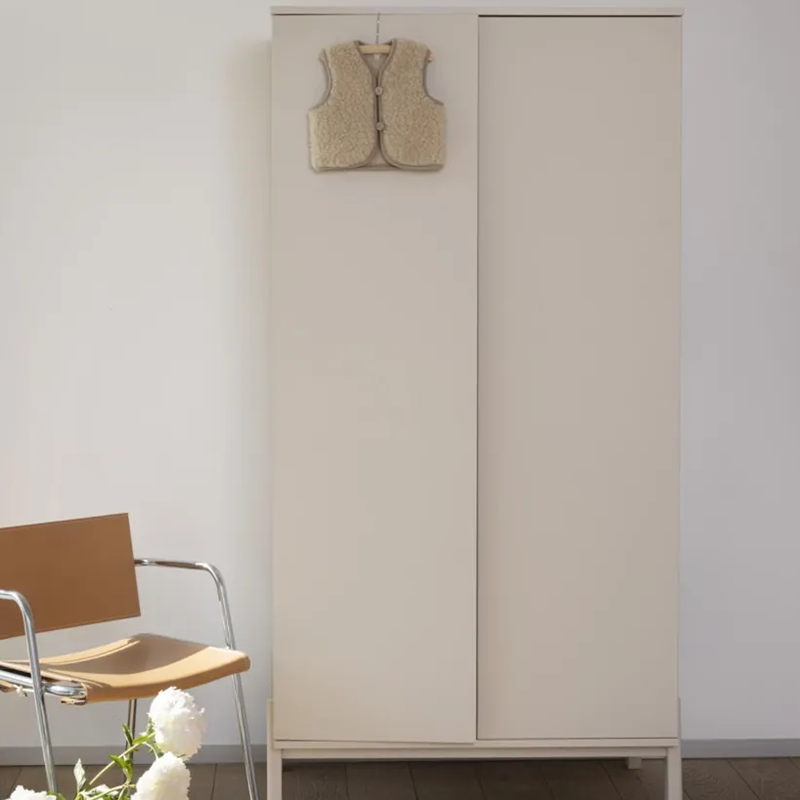 Zweitüriger Schrank mit Füssen lehmfarbrig von Quax steht in einem Zimmer. Mit Babyweste an der Tür und Stuhl neben dem Schrank.