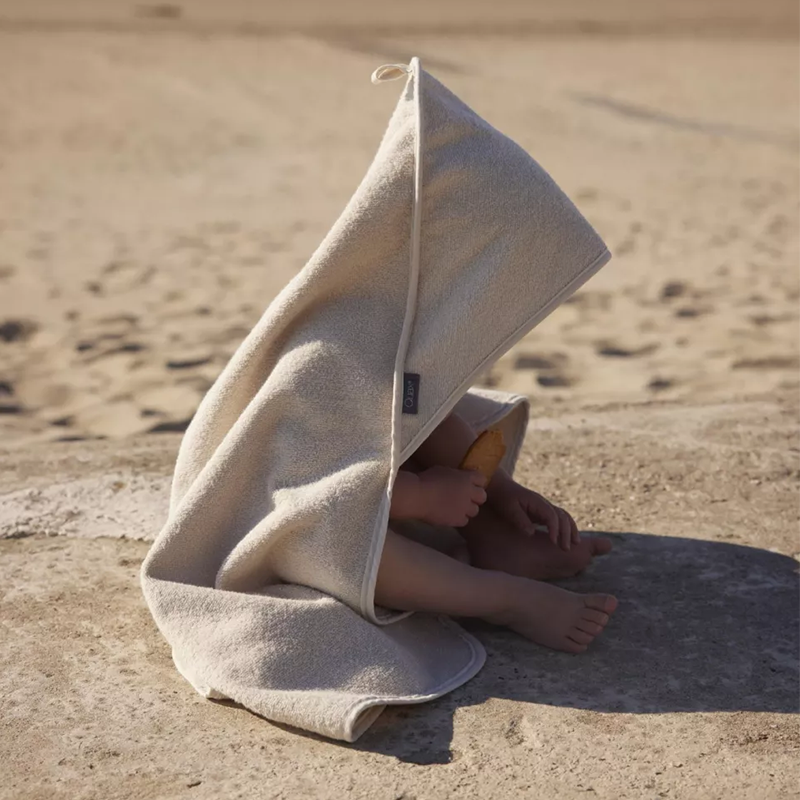 Kind sitzt am Strand und trägt ein Kapuzenbadetuch in Grau von Quax. 