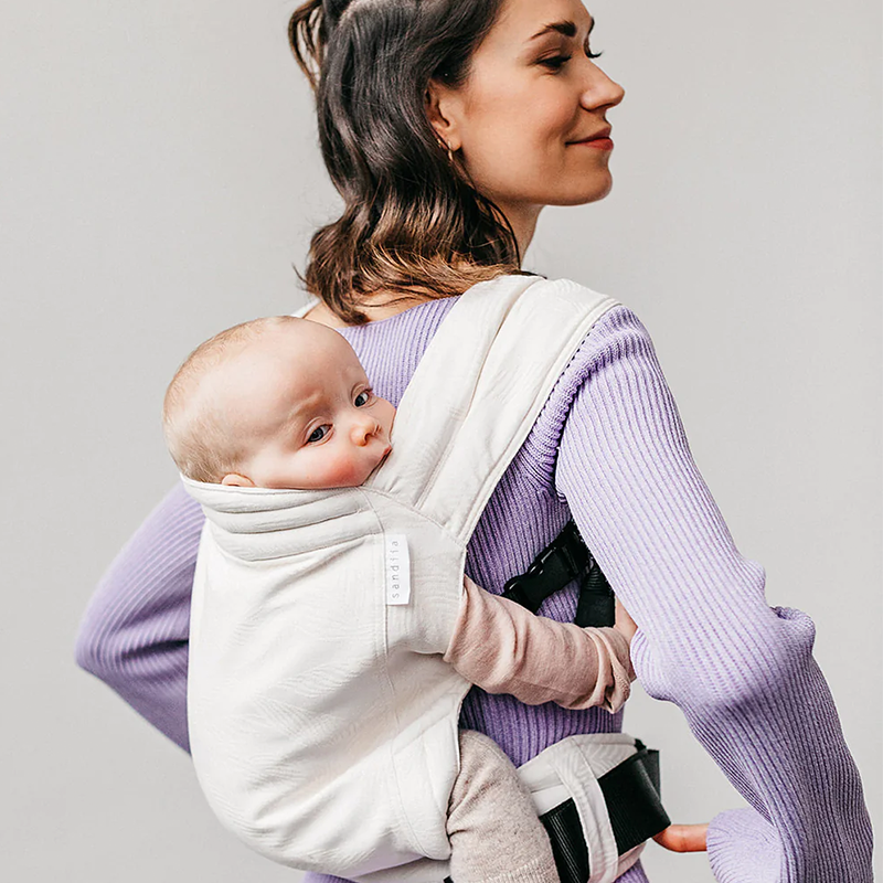Mutter trägt ihr Baby in der Sandiia Babytrage Timeless Palmdiia Pure 2023 auf dem Rücken. 