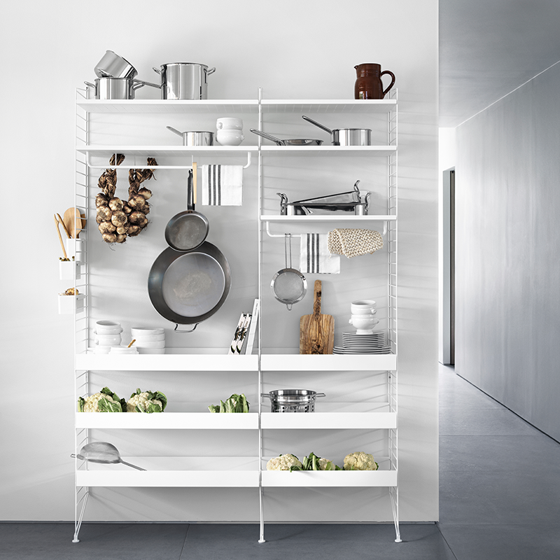 String Furniture Regalsystem in Küche in weiss bestückt mit Töpfen, Schalen, Blumenkohl, Teller und Pfannen. An den Haken hängen Zwiebeln und Pfannen. 