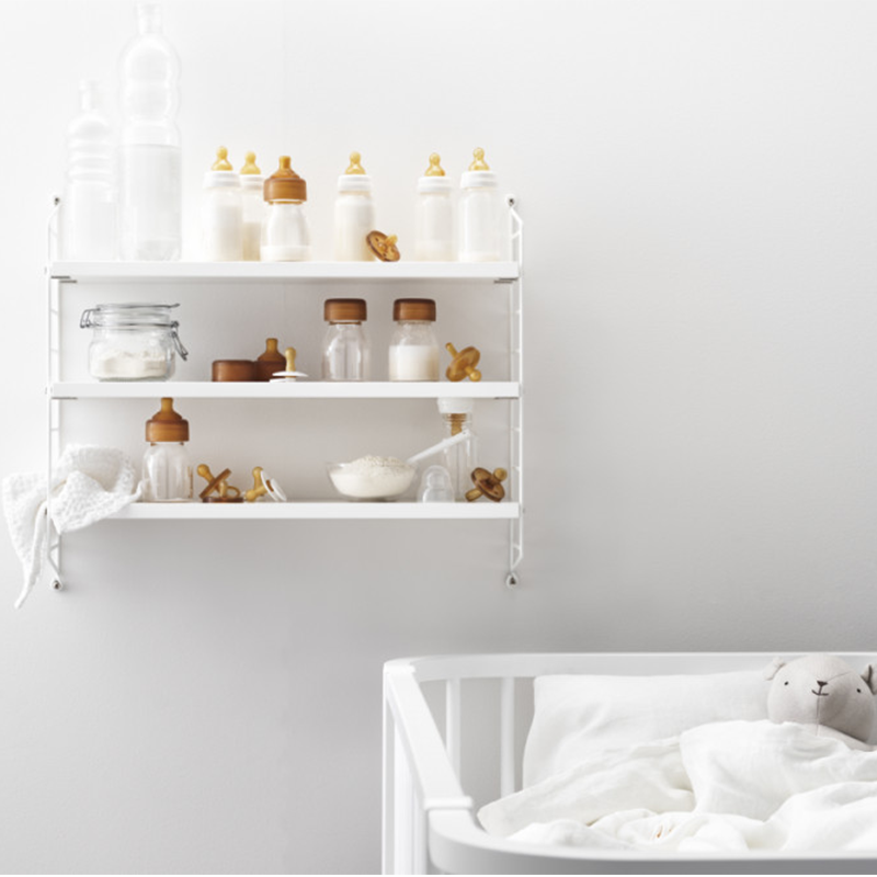 Wandregal Pocket von String in Weiss mit 3 weissen Regalböden hängt oberhalb von einem Babybett. Auf dem Regal hat es Milchpulver und Flaschen und Schnuller. 