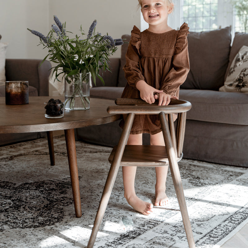 Mädchen steht am Tisch neben dem Oaklings Kinderstuhl &#39;STORM&#39; in Smoked Oak. 